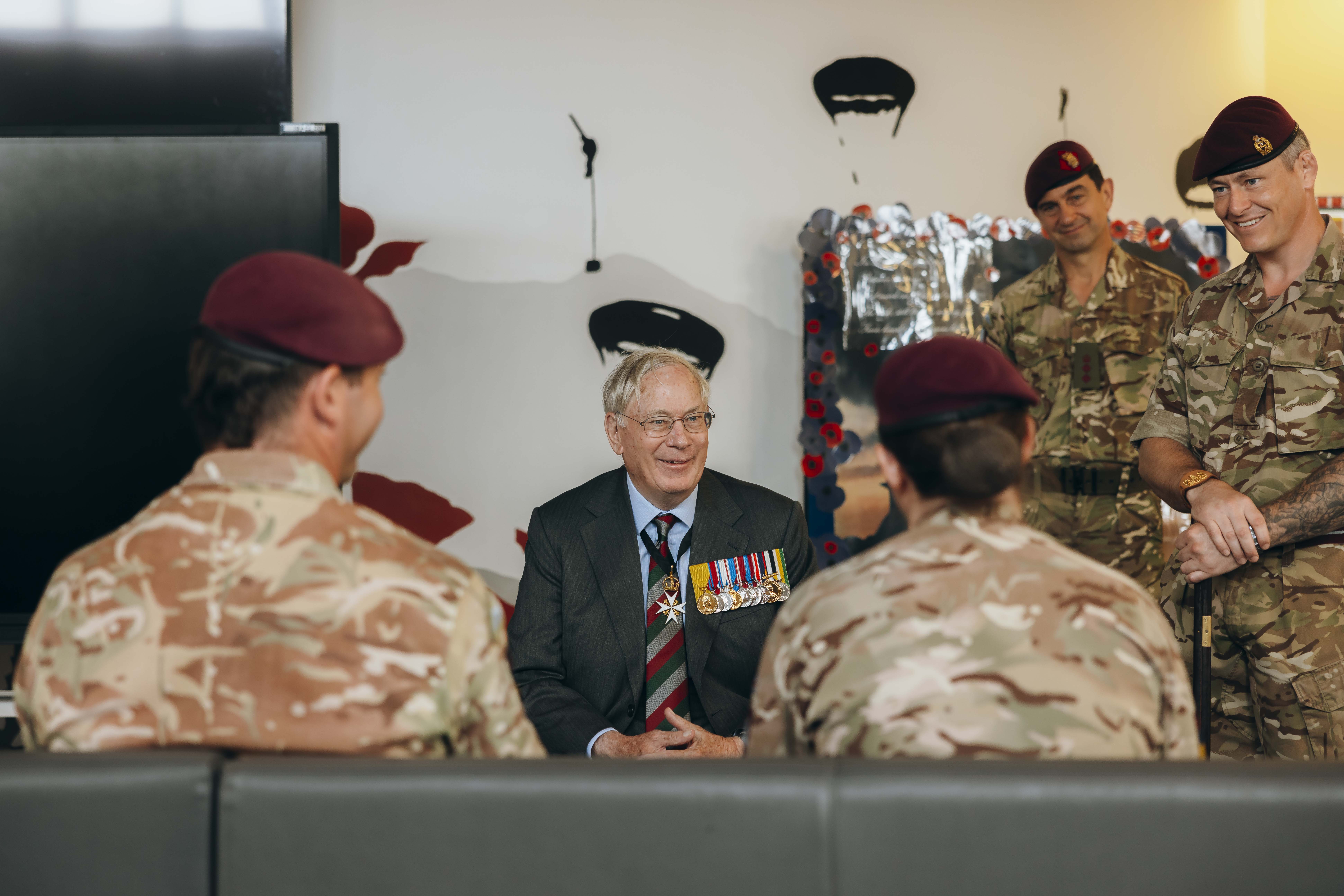 His Royal Highness the Duke of Gloucester sits wearing a suit. He is smiling while talking to two soldiers sat in front of him.