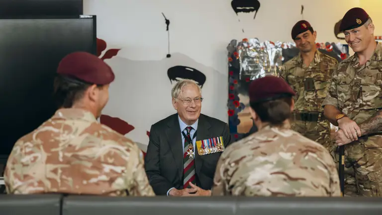His Royal Highness the Duke of Gloucester sits wearing a suit. He is smiling while talking to two soldiers sat in front of him.