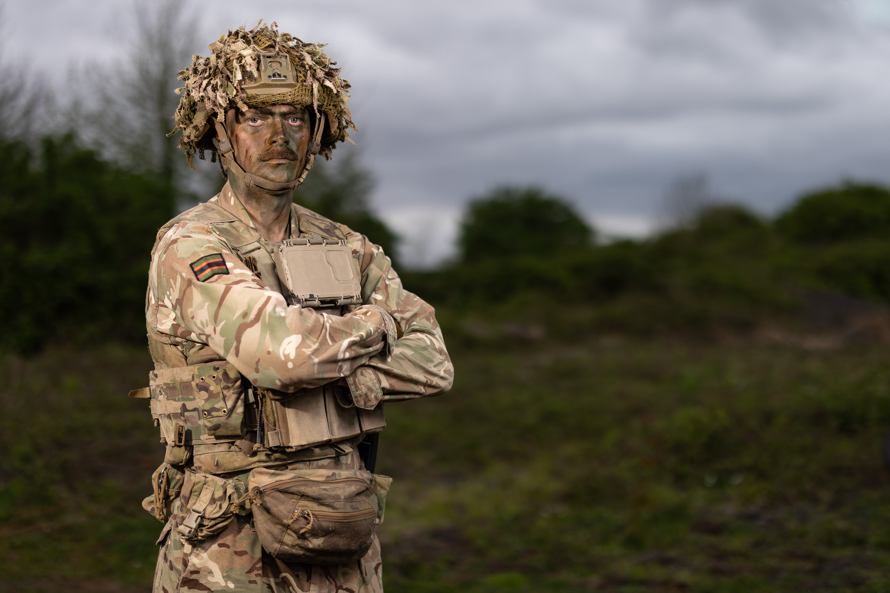 Man stood in camouflage uniform with his arms folded with trees and fields behind him