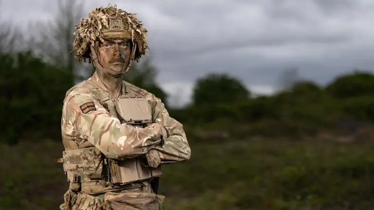 Man stood in camouflage uniform with his arms folded with trees and fields behind him