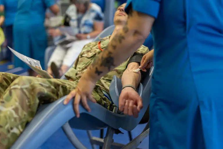 A soldier in camouflage uniform donates blood while a nurse in blue scrubs applies a cotton swab to his arm.