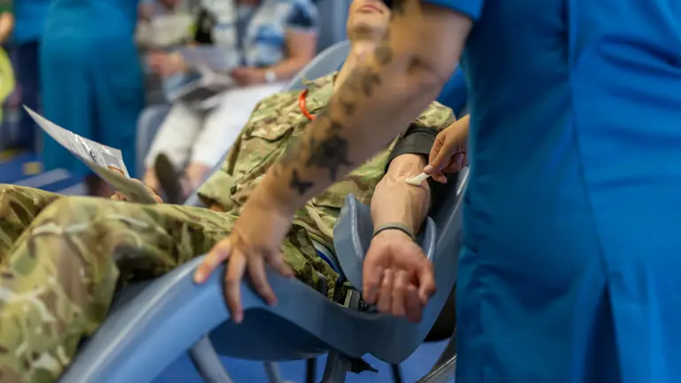 A soldier in camouflage uniform donates blood while a nurse in blue scrubs applies a cotton swab to his arm.