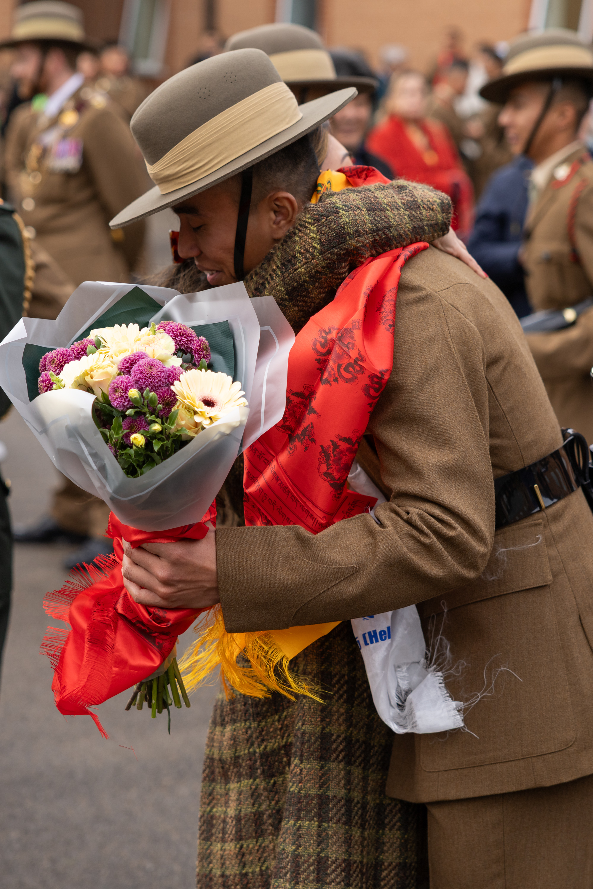  smiling individual in a military uniform, wearing a wide-brimmed hat, holds a colourful bouquet of flowers. The background shows fellow soldiers in uniform