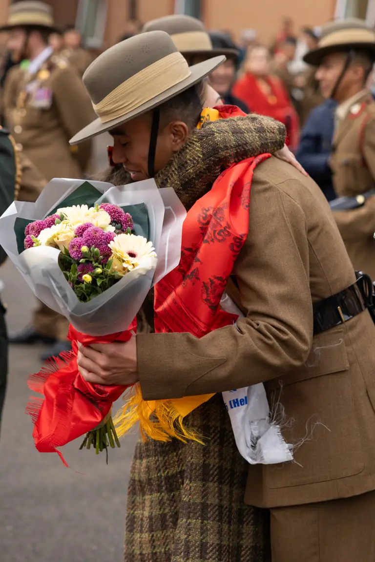 smiling individual in a military uniform, wearing a wide-brimmed hat, holds a colourful bouquet of flowers. The background shows fellow soldiers in uniform