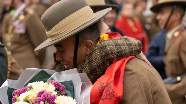 smiling individual in a military uniform, wearing a wide-brimmed hat, holds a colourful bouquet of flowers. The background shows fellow soldiers in uniform