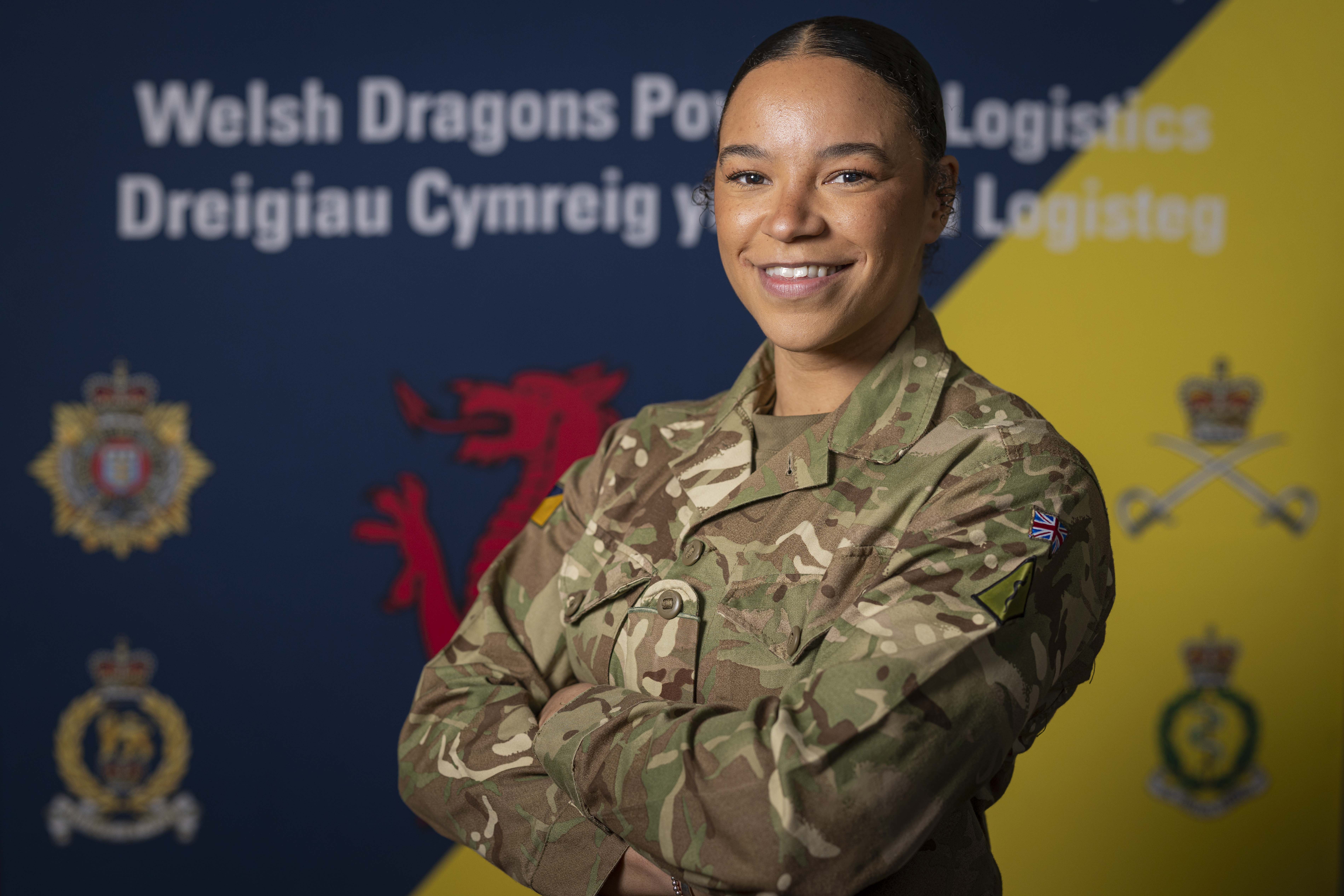 British Army soldier in camouflage uniform stands confidently with arms crossed against a backdrop featuring Welsh and logistics symbols.