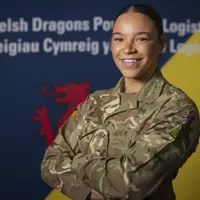 British Army soldier in camouflage uniform stands confidently with arms crossed against a backdrop featuring Welsh and logistics symbols.