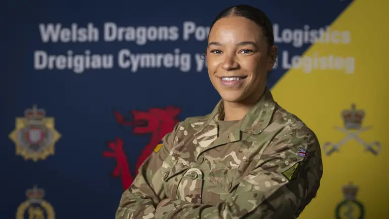 British Army soldier in camouflage uniform stands confidently with arms crossed against a backdrop featuring Welsh and logistics symbols.