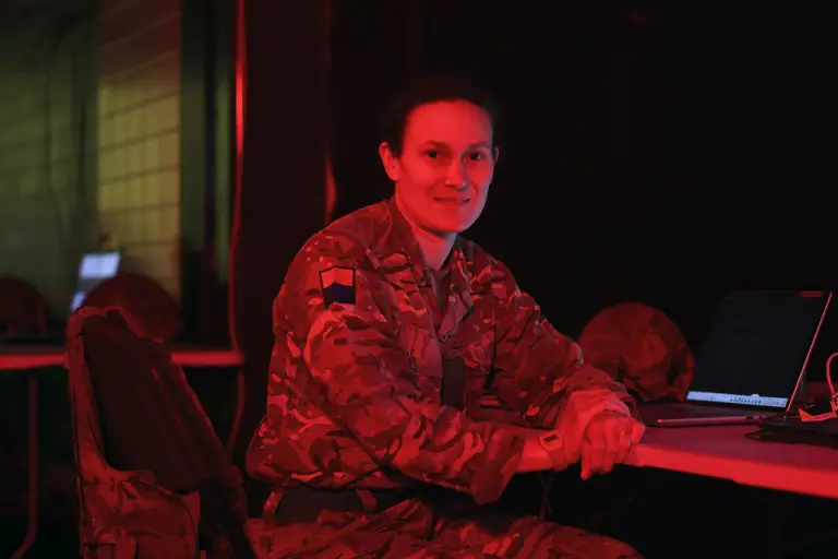 A person in military uniform sits at a table with a laptop, in a room with red lighting. They appear engaged.