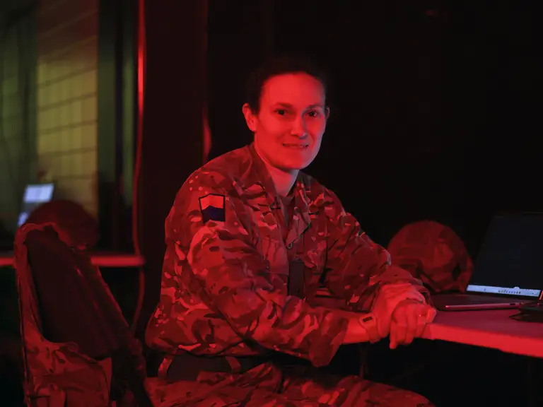 A person in military uniform sits at a table with a laptop, in a room with red lighting. They appear engaged.