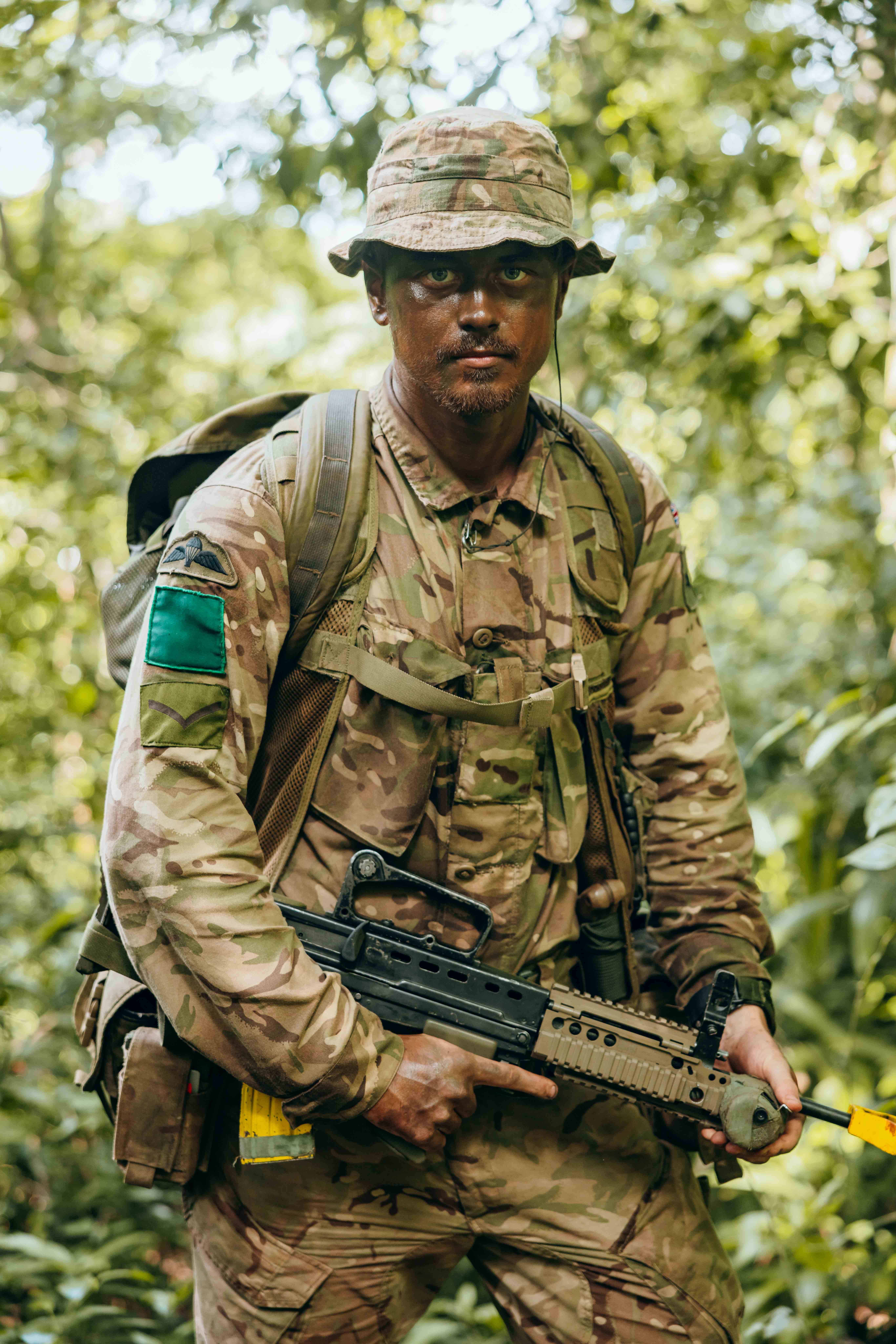 Soldier in full camouflage gear holding a rifle while standing in dense green jungle foliage.