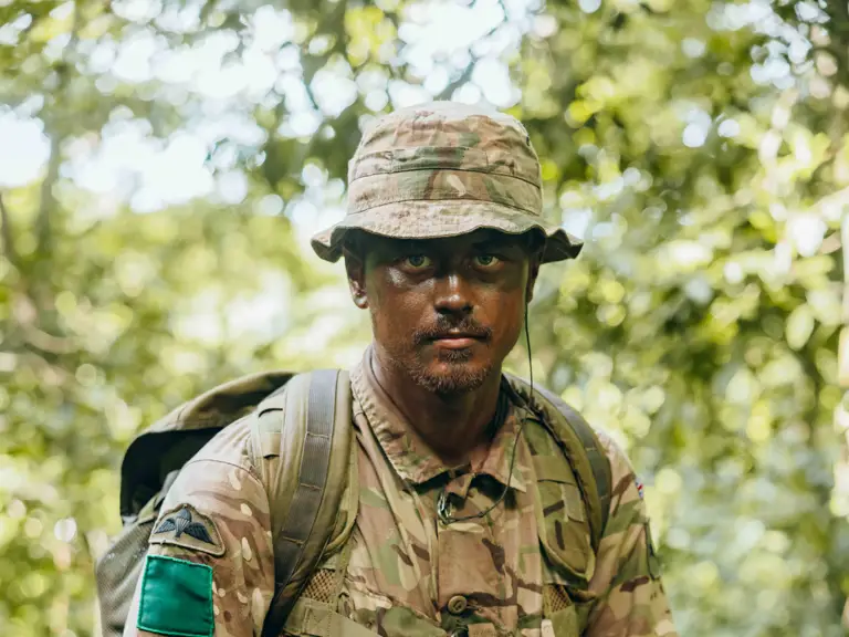 Soldier in full camouflage gear holding a rifle while standing in dense green jungle foliage.