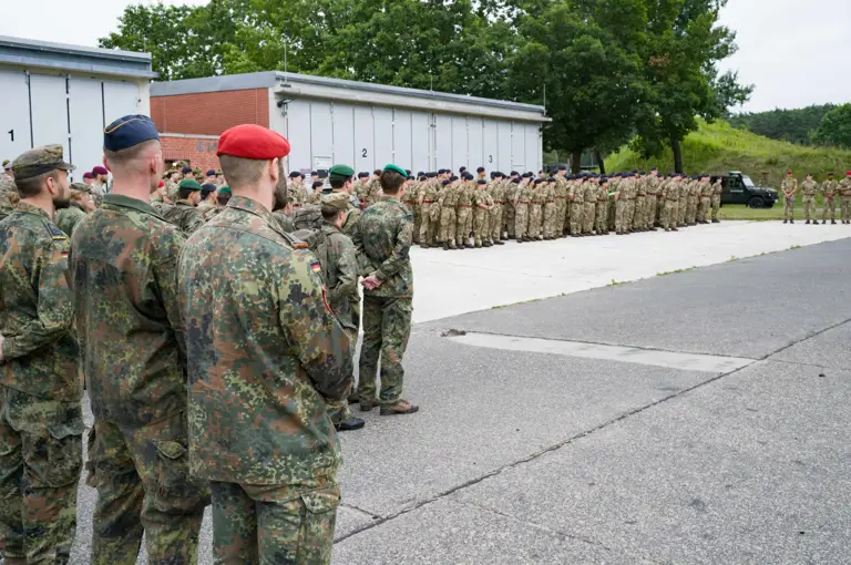A large group of military personnel in camouflage uniforms stand in formation outside, with some wearing colored berets. They are gathered near industrial buildings and surrounded by greenery. A military vehicle is visible in the background.