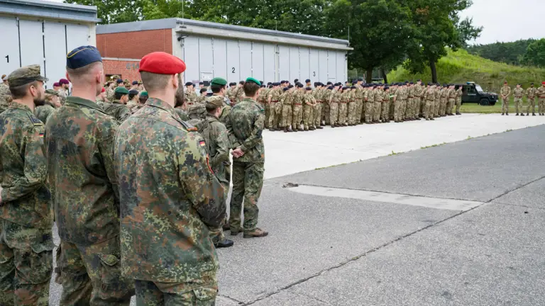 A large group of military personnel in camouflage uniforms stand in formation outside, with some wearing colored berets. They are gathered near industrial buildings and surrounded by greenery. A military vehicle is visible in the background.