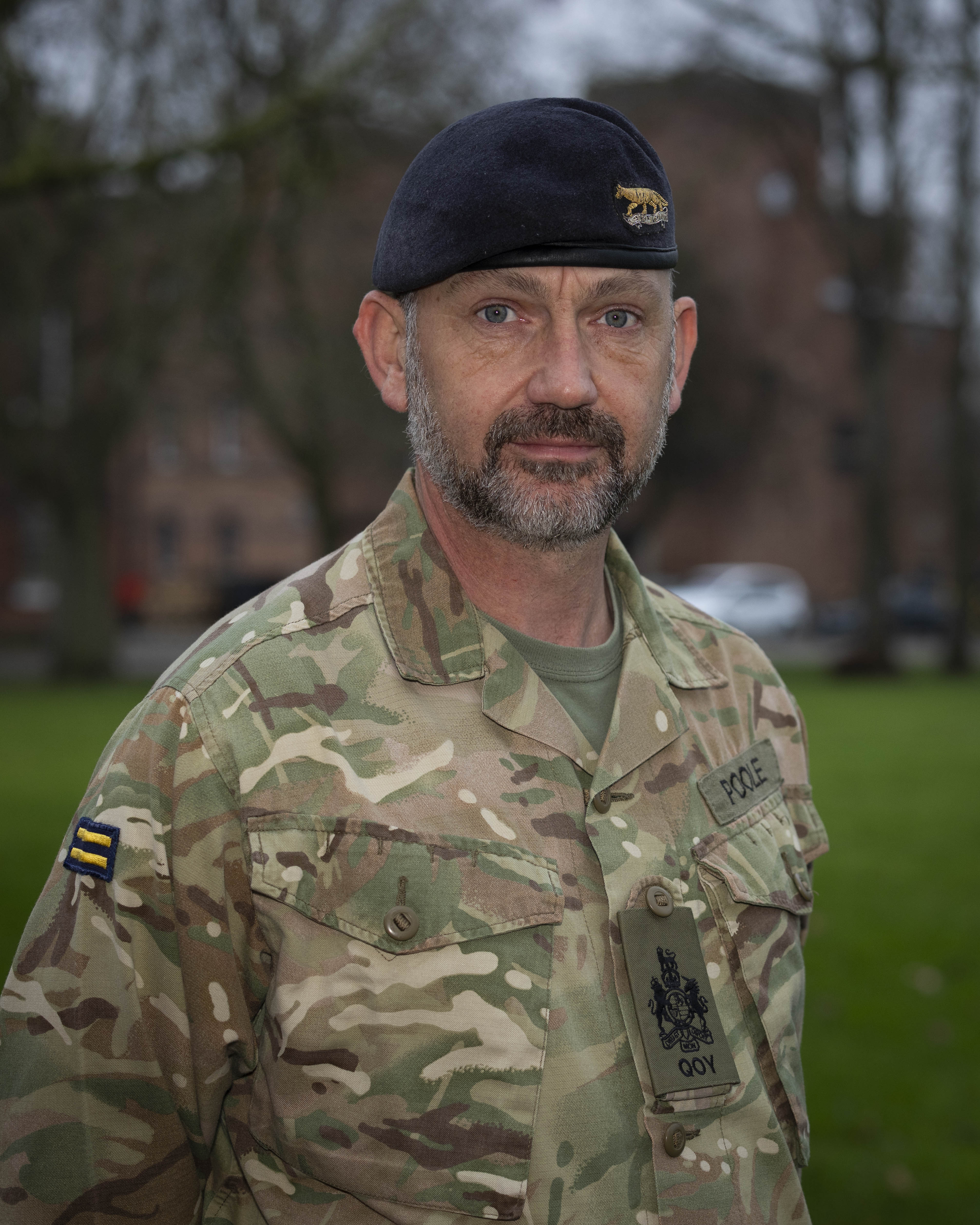 Military personnel in camouflage uniform and dark beret standing outdoors with blurred trees and buildings in the background.