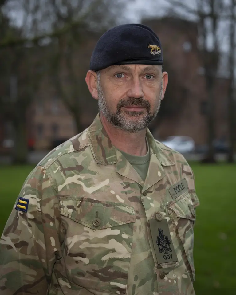 Military personnel in camouflage uniform and dark beret standing outdoors with blurred trees and buildings in the background.