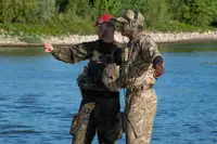 Two soldiers in camouflage uniforms stand by a river, one pointing towards the distance while the other listens.