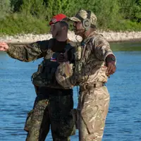Two soldiers in camouflage uniforms stand by a river, one pointing towards the distance while the other listens.