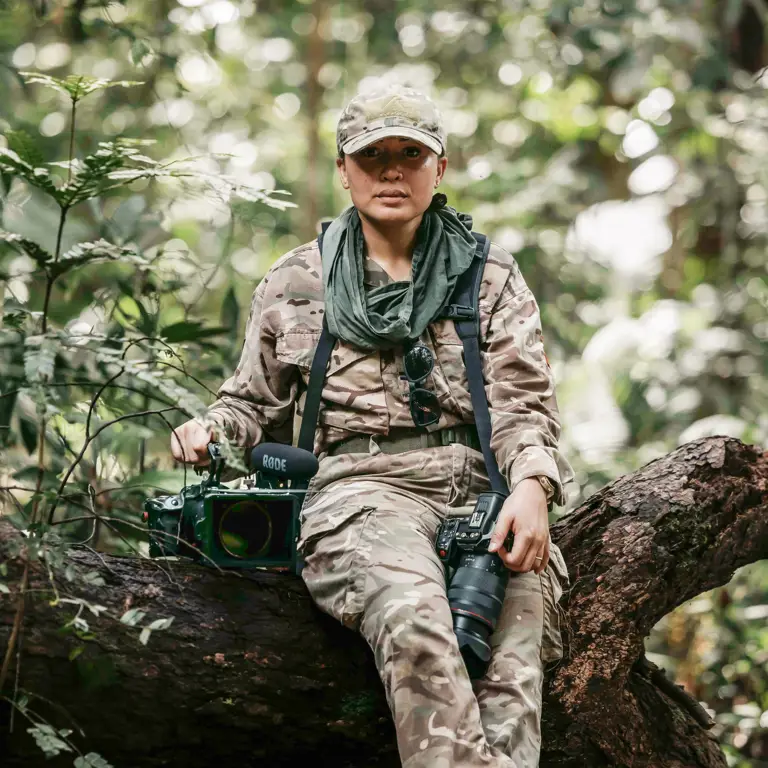 A young female Army Photographer sits on a fallen tree trunk while holding her cameras in the dense forest of Brunei.