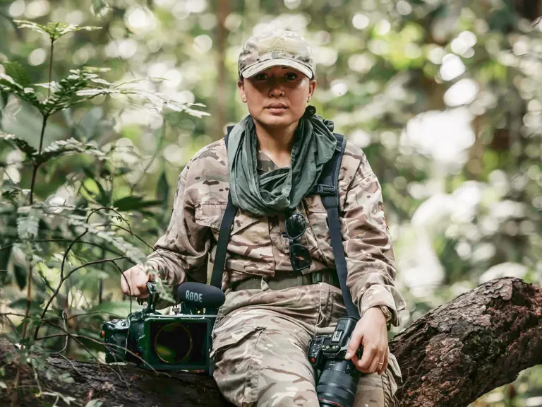 A young female Army Photographer sits on a fallen tree trunk while holding her cameras in the dense forest of Brunei.