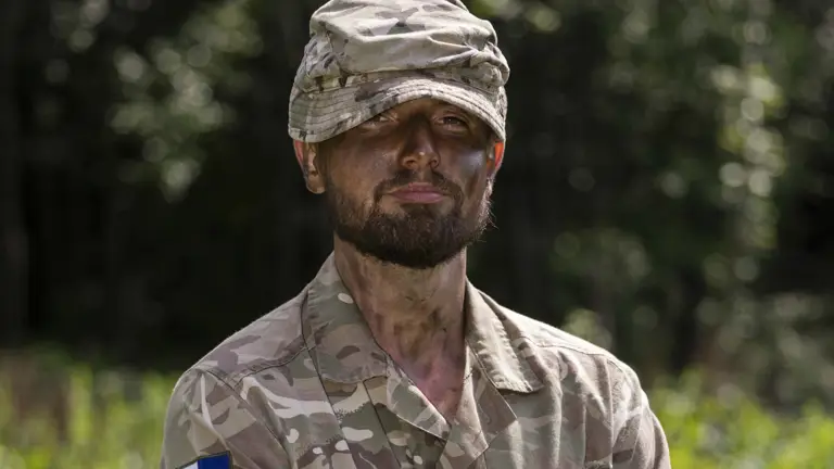 Portrait of soldier in camouflage uniform and hat with a green grassy background.