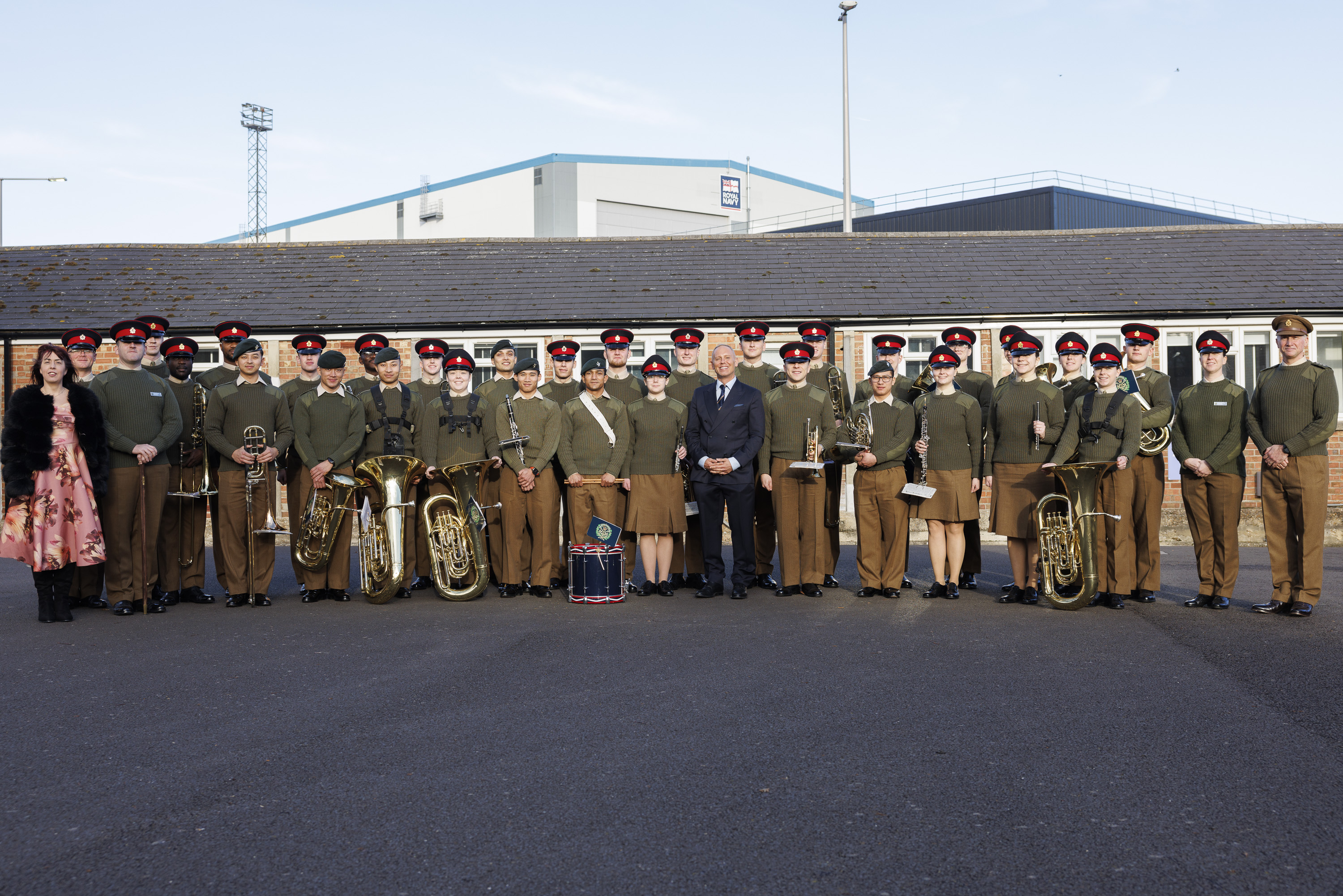 A group photo of uniformed band members and individuals standing outside on a clear day. They hold brass instruments and drums, conveying a formal and proud tone.