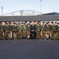 A group photo of uniformed band members and individuals standing outside on a clear day. They hold brass instruments and drums, conveying a formal and proud tone.