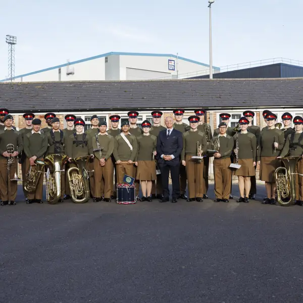 A group photo of uniformed band members and individuals standing outside on a clear day. They hold brass instruments and drums, conveying a formal and proud tone.