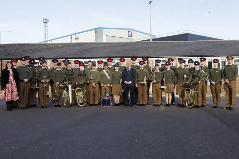 A group photo of uniformed band members and individuals standing outside on a clear day. They hold brass instruments and drums, conveying a formal and proud tone.