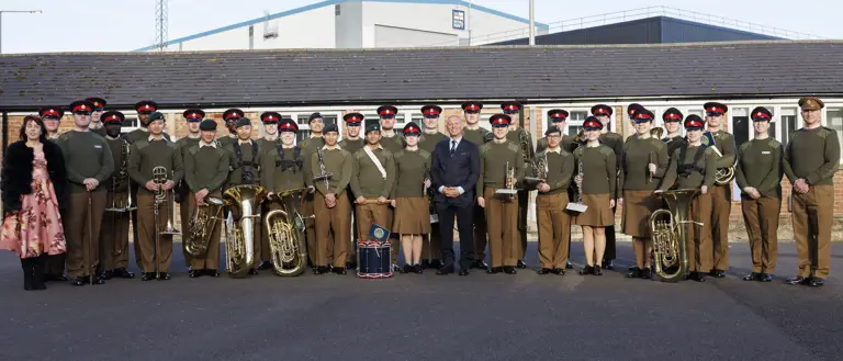A group photo of uniformed band members and individuals standing outside on a clear day. They hold brass instruments and drums, conveying a formal and proud tone.