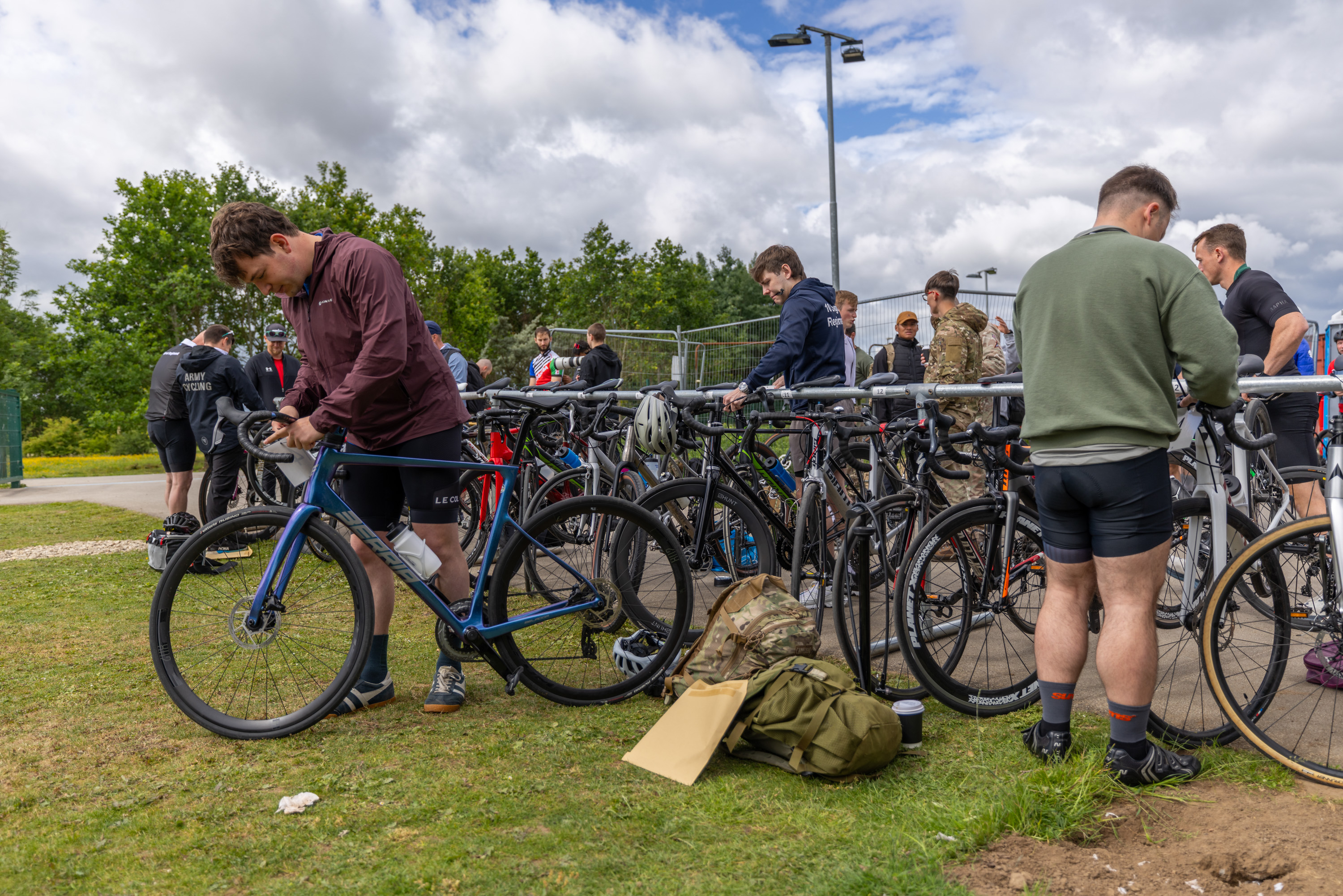 A group of individuals stand near bicycles, some adjusting bikes or gear. A folded green jacket and helmet lie on the grass. People are casually dressed, and a cloudy sky is overhead.