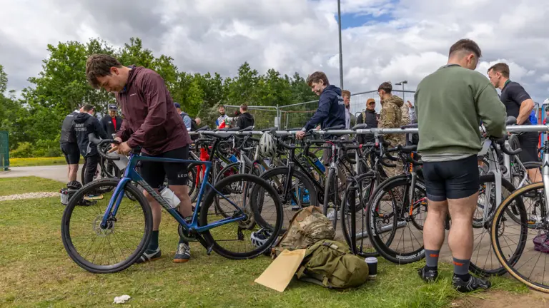 A group of individuals stand near bicycles, some adjusting bikes or gear. A folded green jacket and helmet lie on the grass. People are casually dressed, and a cloudy sky is overhead.