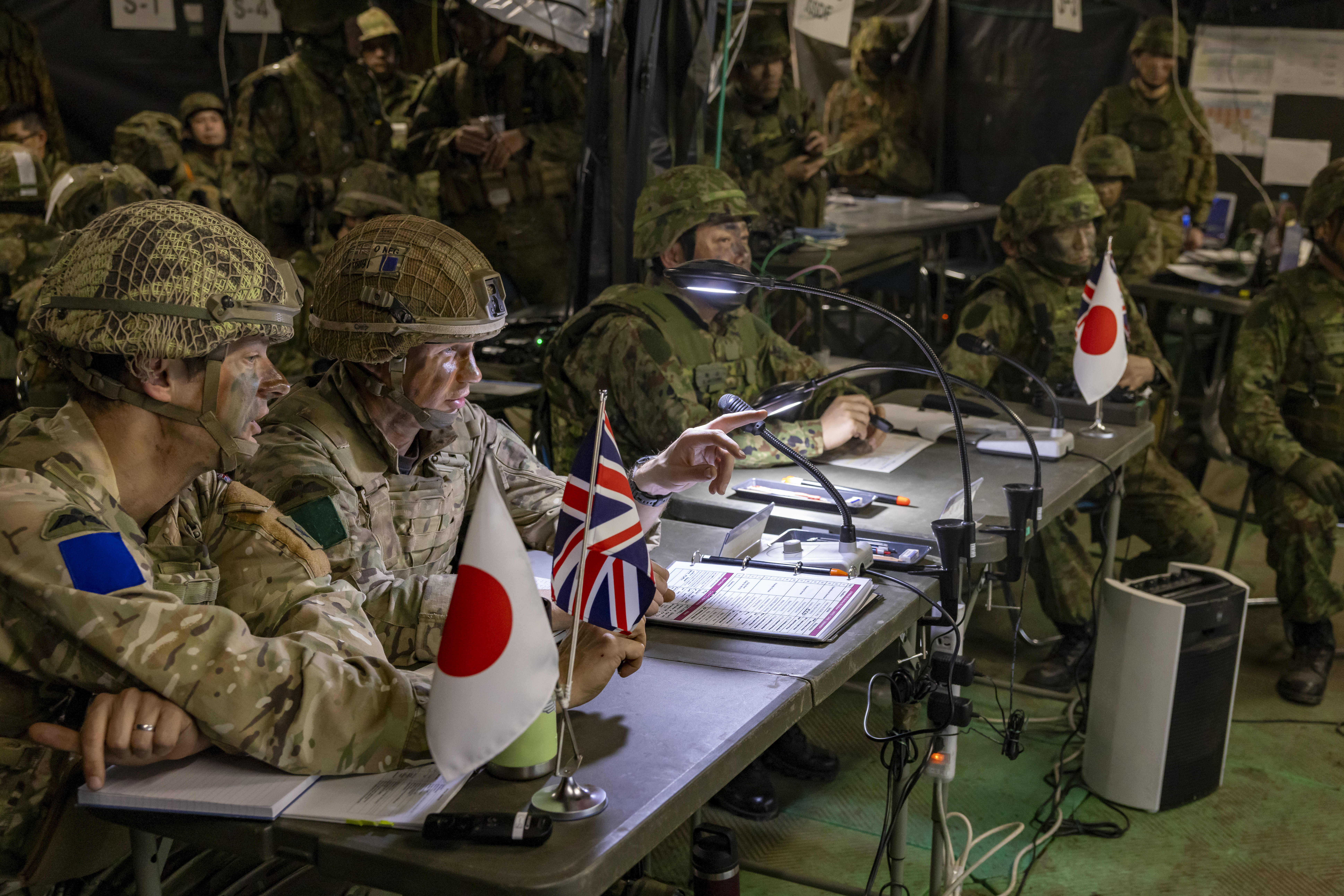 Both British and Japanese soldiers can be seen on uniform sat around field tables to share information and planning to defeat the enemy whilst on exercise in japan. Both Union Jack and Japanese flags can be seen on the tables along with notepads, microphones and lights clamped onto the tables.