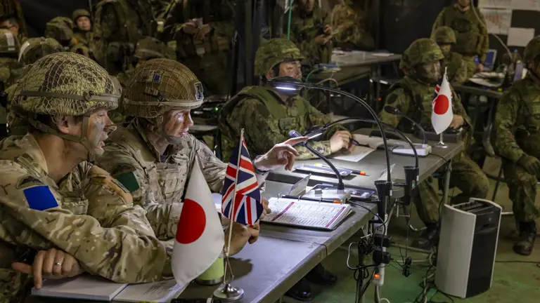 Both British and Japanese soldiers can be seen on uniform sat around field tables to share information and planning to defeat the enemy whilst on exercise in japan. Both Union Jack and Japanese flags can be seen on the tables along with notepads, microphones and lights clamped onto the tables.