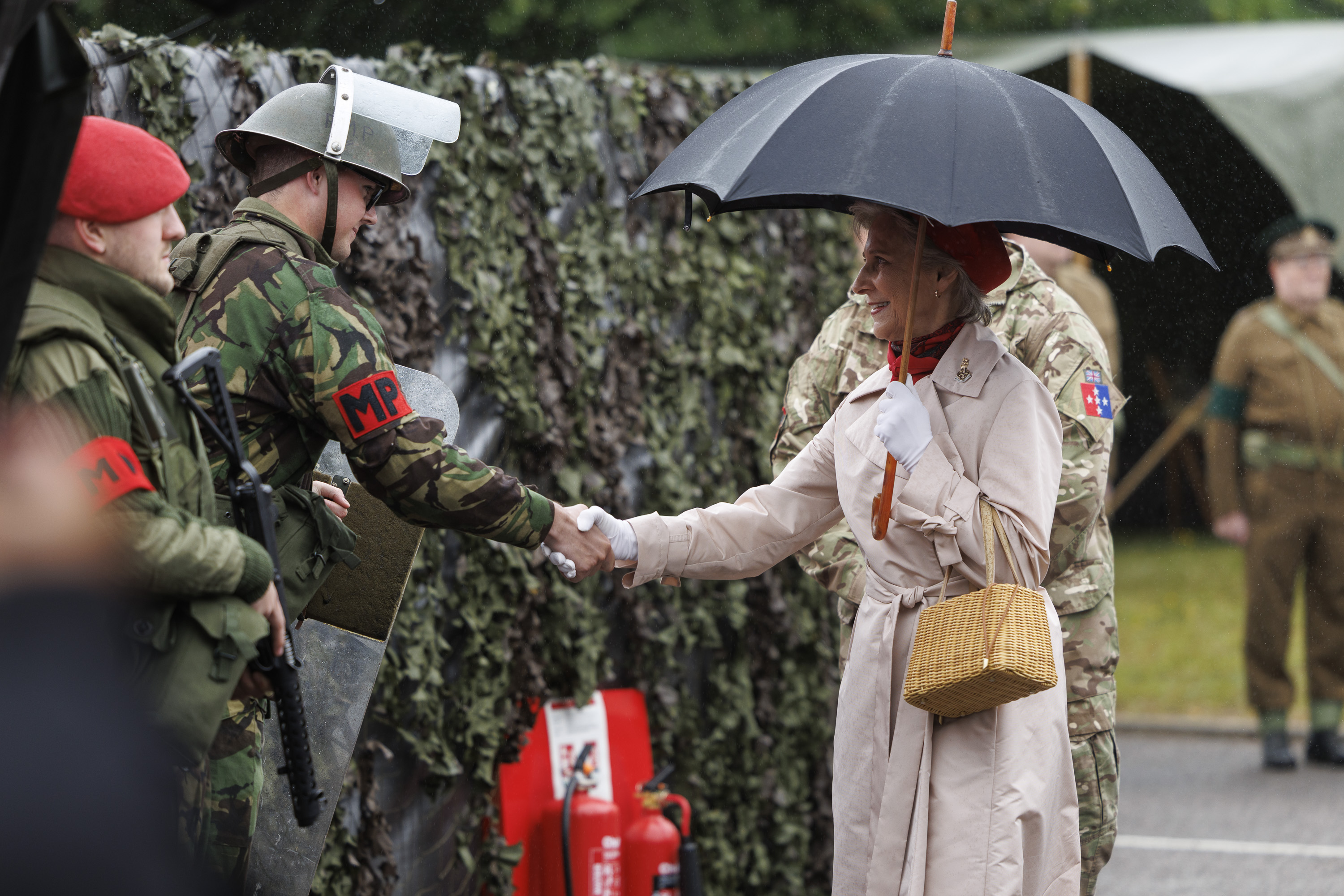 Women in cream coat and red hat now carrying an umbrella shakes hand of soldiers in camouflage uniform with a red MP patch on their arm and wearing a metal green hat 