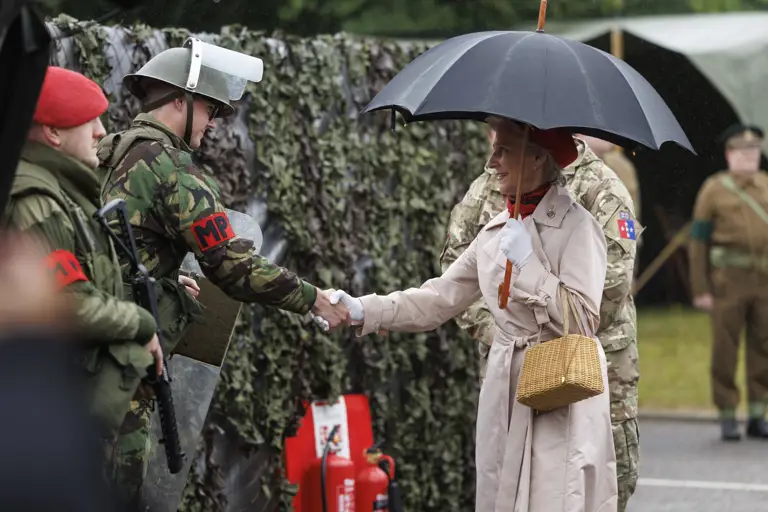 Women in cream coat and red hat now carrying an umbrella shakes hand of soldiers in camouflage uniform with a red MP patch on their arm and wearing a metal green hat