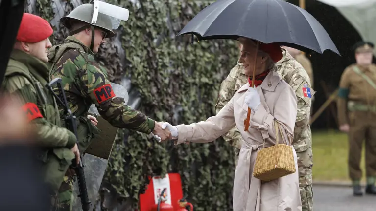 Women in cream coat and red hat now carrying an umbrella shakes hand of soldiers in camouflage uniform with a red MP patch on their arm and wearing a metal green hat