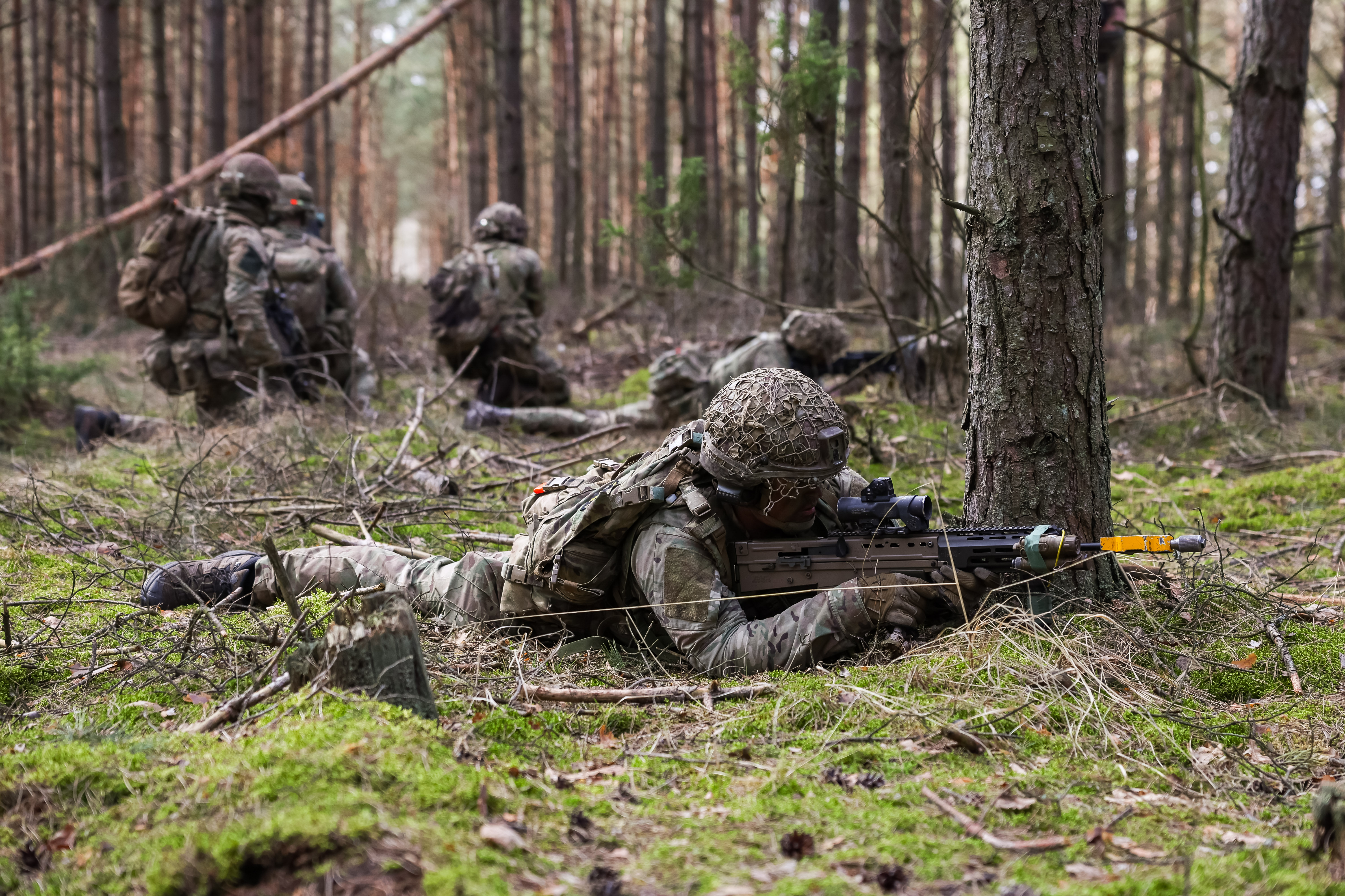 Soldiers lay down taking aim with their rifles. Behind them, three soldiers kneel together talking.