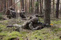 Soldiers lay down taking aim with their rifles. Behind them, three soldiers kneel together talking.