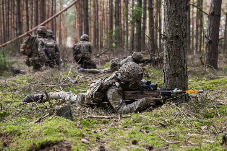 Soldiers lay down taking aim with their rifles. Behind them, three soldiers kneel together talking.