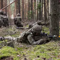 Soldiers lay down taking aim with their rifles. Behind them, three soldiers kneel together talking.