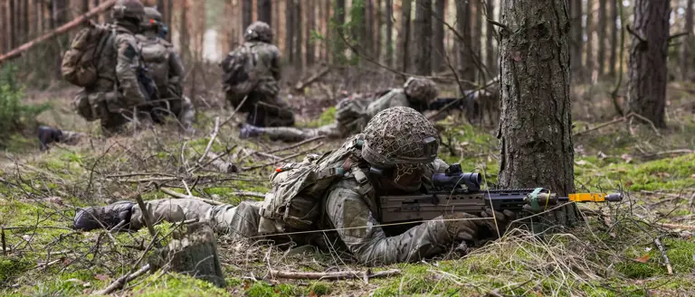 Soldiers lay down taking aim with their rifles. Behind them, three soldiers kneel together talking.