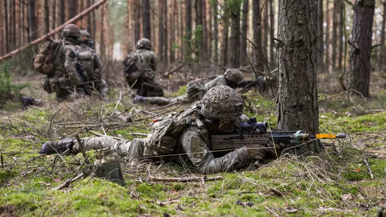 Soldiers lay down taking aim with their rifles. Behind them, three soldiers kneel together talking.