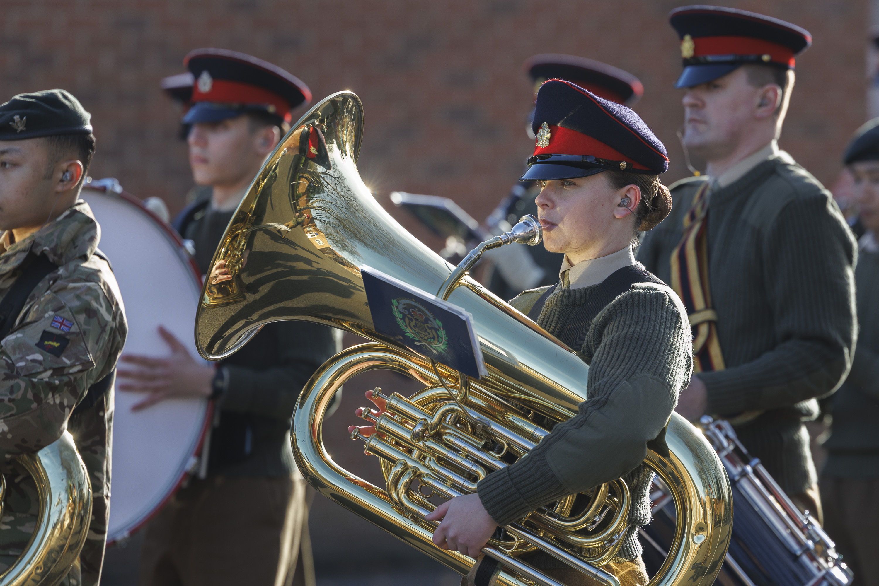 Military band members in uniform perform outdoors. A prominent tuba player in a peaked cap is in focus. The scene conveys discipline and focus.