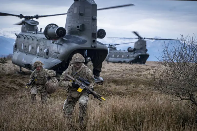 Two soldiers in camouflage uniform are seen running out the back of a Chinook with rifles and army gear into a field.