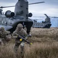 Two soldiers in camouflage uniform are seen running out the back of a Chinook with rifles and army gear into a field.