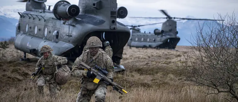 Two soldiers in camouflage uniform are seen running out the back of a Chinook with rifles and army gear into a field.