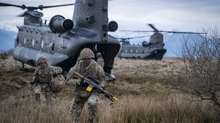 Two soldiers in camouflage uniform are seen running out the back of a Chinook with rifles and army gear into a field.