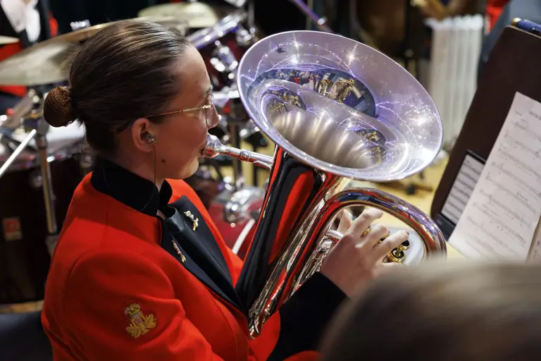 Musician in a red uniform playing a shiny brass euphonium during a band performance with sheet music visible.