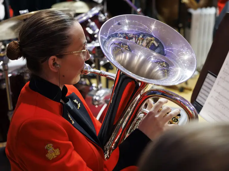 Musician in a red uniform playing a shiny brass euphonium during a band performance with sheet music visible.